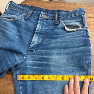 Pu&ograve; includere: Primo piano di jeans blu in denim con un classico design a cinque tasche. Un metro a nastro giallo mostra una lunghezza di circa 25 cm. I jeans hanno una chiusura con bottoni e passanti per cintura.