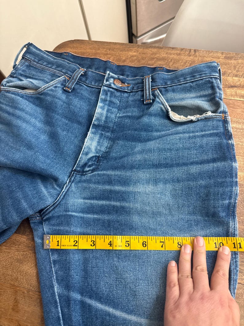 Pu&ograve; includere: Primo piano di jeans blu in denim con un classico design a cinque tasche. Un metro a nastro giallo mostra una lunghezza di circa 25 cm. I jeans hanno una chiusura con bottoni e passanti per cintura.