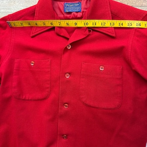 以下が含まれることがあります： 鮮やかな赤色のボタンダウンシャツ。胸ポケットが2つと襟が付いています。黄色のメジャーで計測すると、長さは約40cmです。Pendletonのラベルが見えます。