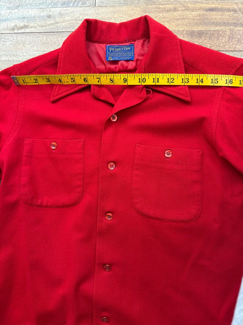 以下が含まれることがあります： 鮮やかな赤色のボタンダウンシャツ。胸ポケットが2つと襟が付いています。黄色のメジャーで計測すると、長さは約40cmです。Pendletonのラベルが見えます。