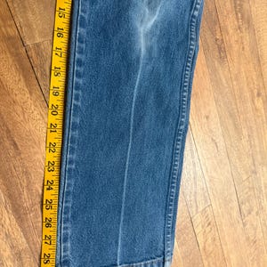 May include: Close-up of blue denim jeans with a yellow measuring tape alongside. The tape shows measurements from 12 to 31 inches. The jeans have a faded appearance and a vertical seam down the leg.