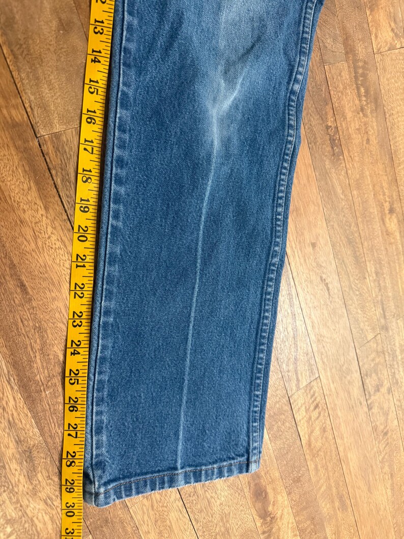 May include: Close-up of blue denim jeans with a yellow measuring tape alongside. The tape shows measurements from 12 to 31 inches. The jeans have a faded appearance and a vertical seam down the leg.