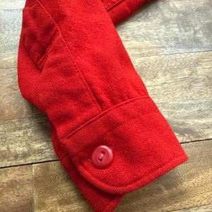 以下が含まれることがあります： 鮮やかな赤色のジャケットの袖のクローズアップ。 袖にはボタン付きのカフスと小さなポケットが付いています。 生地は質感のある外観です。 ジャケットは、ブレザーやコートなどの大きな衣服の一部である可能性があります。