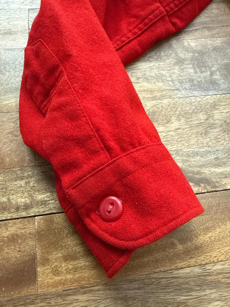 以下が含まれることがあります： 鮮やかな赤色のジャケットの袖のクローズアップ。 袖にはボタン付きのカフスと小さなポケットが付いています。 生地は質感のある外観です。 ジャケットは、ブレザーやコートなどの大きな衣服の一部である可能性があります。