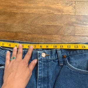 May include: Close-up of blue denim jeans with a yellow measuring tape stretched across the waist. The waist measures approximately 15 inches across. The jeans have a button closure and classic five-pocket design.