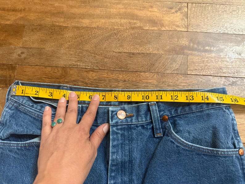 May include: Close-up of blue denim jeans with a yellow measuring tape stretched across the waist. The waist measures approximately 15 inches across. The jeans have a button closure and classic five-pocket design.