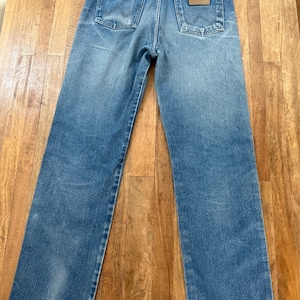 K&ouml;nnte beinhalten: Blaue Jeans aus Denim mit klassischem F&uuml;nf-Taschen-Design. Die Jeans haben eine verwaschene Optik und einen braunen Lederaufn&auml;her auf der Ges&auml;&szlig;tasche. Die Marke Wrangler ist sichtbar.