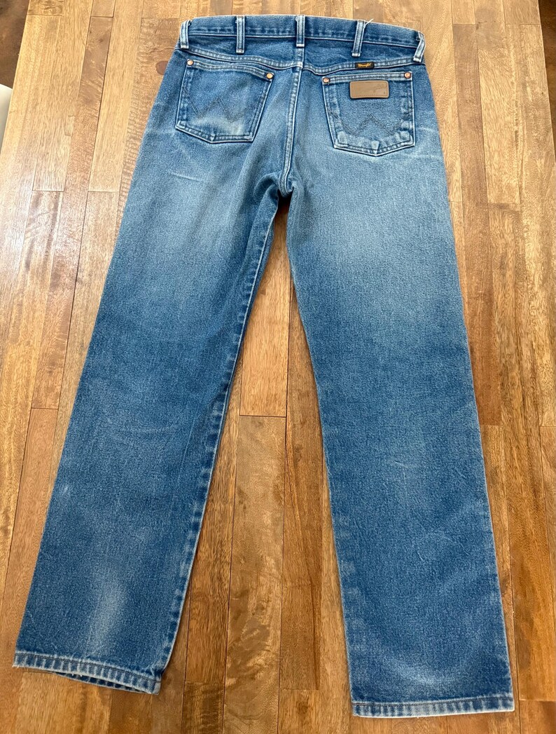 K&ouml;nnte beinhalten: Blaue Jeans aus Denim mit klassischem F&uuml;nf-Taschen-Design. Die Jeans haben eine verwaschene Optik und einen braunen Lederaufn&auml;her auf der Ges&auml;&szlig;tasche. Die Marke Wrangler ist sichtbar.
