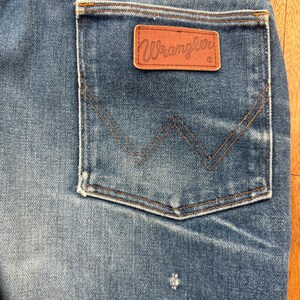 Pu&ograve; includere: Primo piano di jeans di denim blu con un'etichetta Wrangler sulla tasca posteriore. I jeans hanno un classico design a cinque tasche con una toppa in pelle marrone e il logo Wrangler. Le cuciture sono di colore marrone a contrasto.