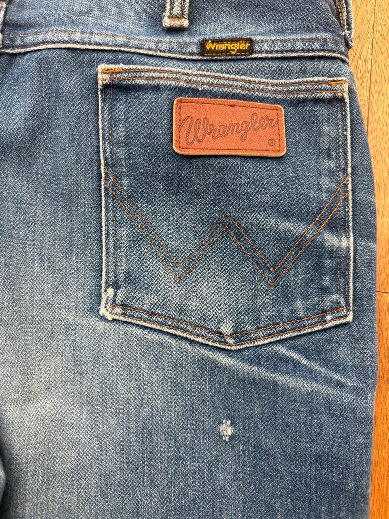 Pu&ograve; includere: Primo piano di jeans di denim blu con un'etichetta Wrangler sulla tasca posteriore. I jeans hanno un classico design a cinque tasche con una toppa in pelle marrone e il logo Wrangler. Le cuciture sono di colore marrone a contrasto.