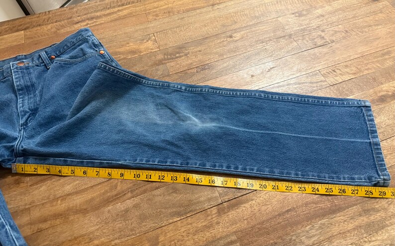 May include: Blue denim jeans with a yellow measuring tape laid across the leg. The jeans are folded and appear to be a size 34 waist. The measuring tape shows a length of approximately 30 inches.