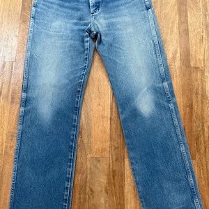 K&ouml;nnte beinhalten: Blaue Jeans aus Denim mit klassischem F&uuml;nf-Taschen-Design. Die Jeans haben eine verwaschene Optik und eine gerade Beinpassform. Das Bild zeigt die Jeans flach auf einer Holzoberfl&auml;che, die die Details des Denims zeigt.