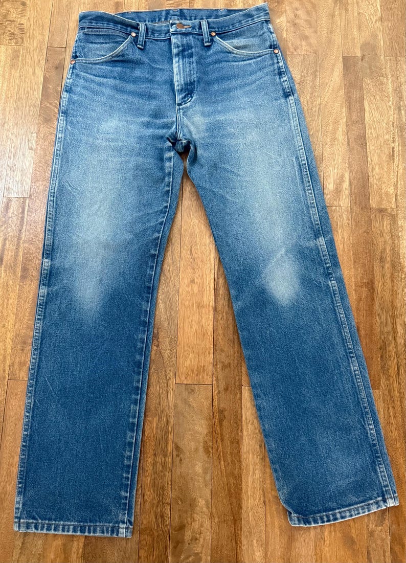 K&ouml;nnte beinhalten: Blaue Jeans aus Denim mit klassischem F&uuml;nf-Taschen-Design. Die Jeans haben eine verwaschene Optik und eine gerade Beinpassform. Das Bild zeigt die Jeans flach auf einer Holzoberfl&auml;che, die die Details des Denims zeigt.