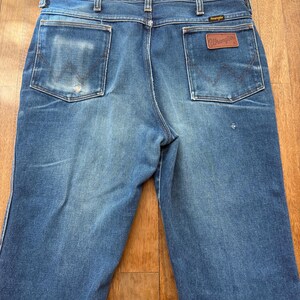 Pu&ograve; includere: Jeans Wrangler blu con un aspetto sbiadito. Le tasche posteriori hanno un design classico e una toppa in pelle marrone con il logo Wrangler &egrave; visibile. I jeans sono su una superficie di legno.