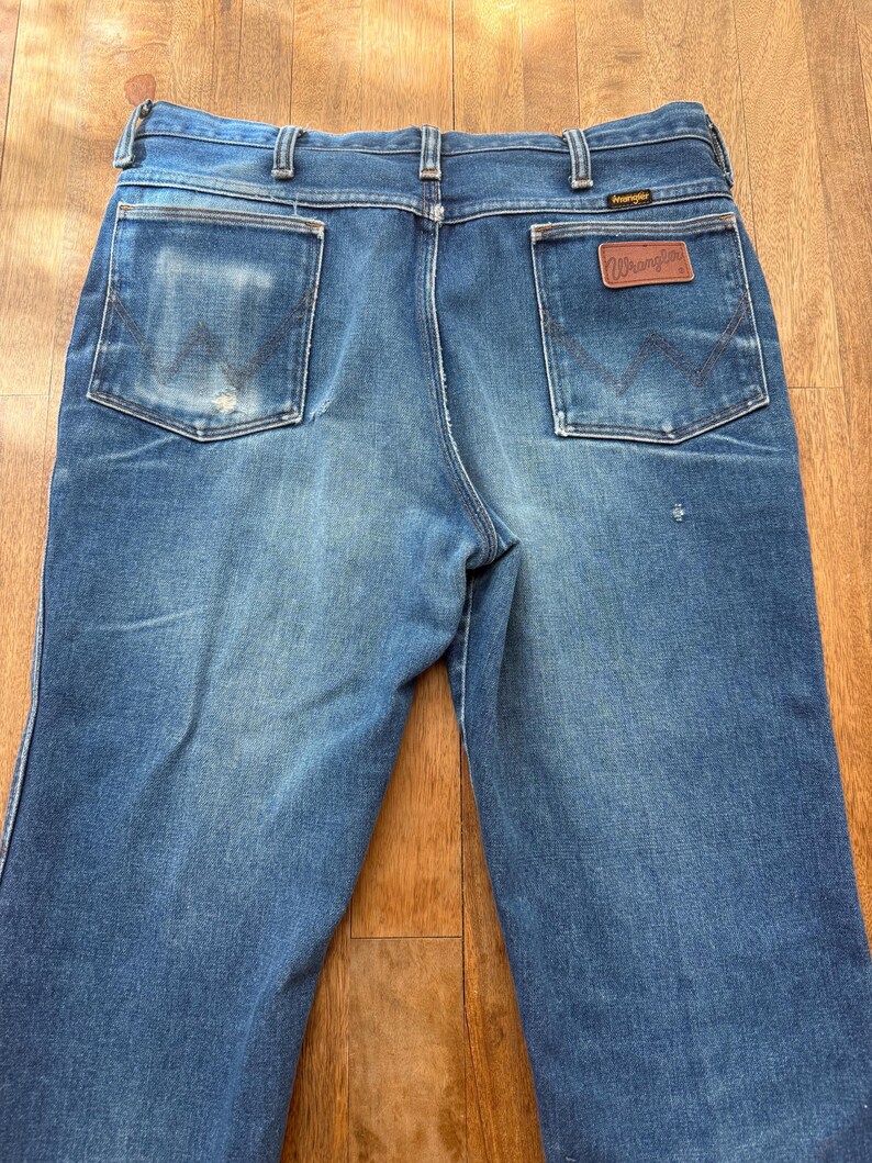 Pu&ograve; includere: Jeans Wrangler blu con un aspetto sbiadito. Le tasche posteriori hanno un design classico e una toppa in pelle marrone con il logo Wrangler &egrave; visibile. I jeans sono su una superficie di legno.