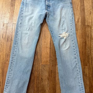 May include: Light blue denim jeans featuring a classic five-pocket design, a button fly closure, and a distressed finish with a prominent frayed rip on the left knee area. The fabric displays a faded wash with whiskering details at the hips.