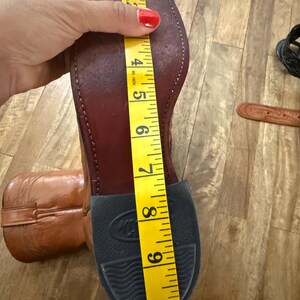 May include: A brown leather cowboy boot with a black rubber sole. A yellow measuring tape shows the boot's length is approximately 10 inches. The boot is on a wooden surface.