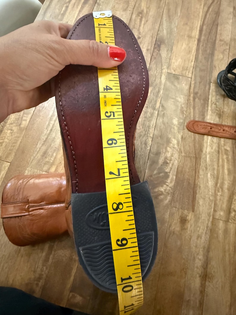 May include: A brown leather cowboy boot with a black rubber sole. A yellow measuring tape shows the boot's length is approximately 10 inches. The boot is on a wooden surface.