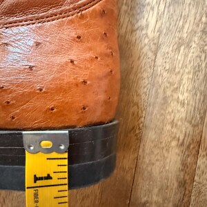 May include: Close-up of a brown leather cowboy boot with a textured surface. The heel measures approximately 1 inch. The boot is set against a wooden background, with a yellow measuring tape.