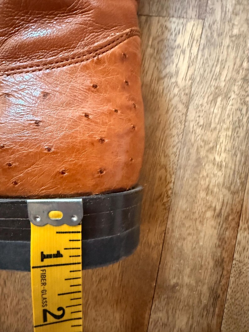 May include: Close-up of a brown leather cowboy boot with a textured surface. The heel measures approximately 1 inch. The boot is set against a wooden background, with a yellow measuring tape.