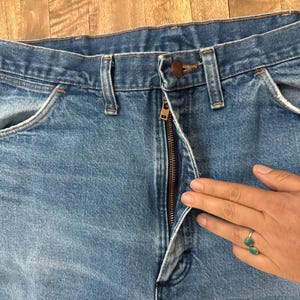 May include: Close-up of light blue denim jeans with a partially unzipped bronze zipper. The jeans have classic five-pocket styling, including front pockets and a coin pocket. The jeans are on a wooden surface.