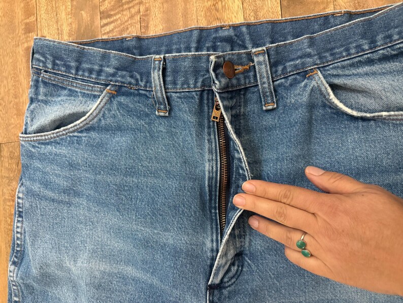 May include: Close-up of light blue denim jeans with a partially unzipped bronze zipper. The jeans have classic five-pocket styling, including front pockets and a coin pocket. The jeans are on a wooden surface.