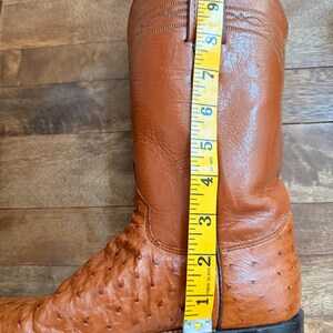 May include: A brown leather cowboy boot with a textured ostrich leg and a smooth upper. The boot is measured with a yellow tape measure, showing a height of approximately 10 inches.