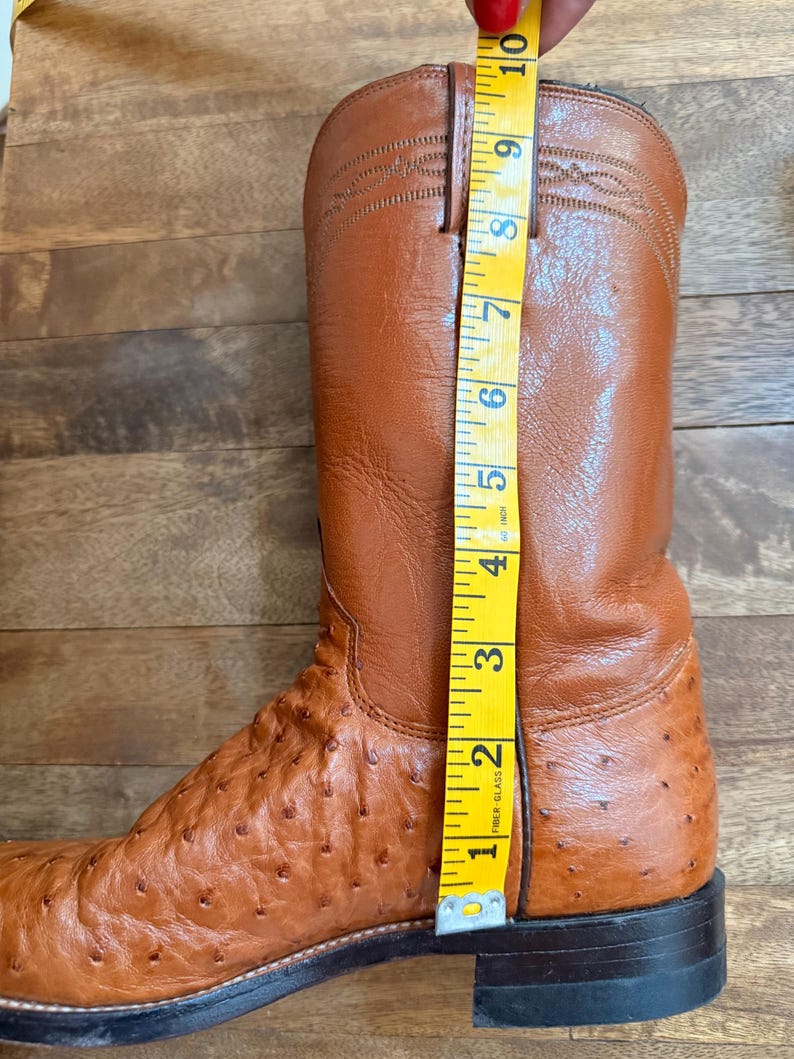 May include: A brown leather cowboy boot with a textured ostrich leg and a smooth upper. The boot is measured with a yellow tape measure, showing a height of approximately 10 inches.