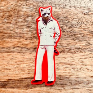 May include: A die-cut sticker featuring a raccoon's head on a person's body, wearing a white suit with a collared shirt and flared pants. The sticker has a red border and is set against a wood grain background.