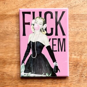 May include: A rectangular pink magnet with the words "FUCK 'EM" in black. The magnet features a vintage illustration of a woman in a black dress blowing a bubblegum bubble. She wears a pearl necklace, gloves, and has flowers in her hair.