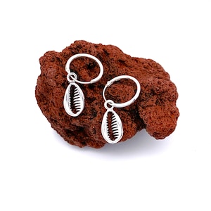 May include: A pair of silver hoop earrings with a shell charm hanging from each hoop. The earrings are displayed on a brown rock.
