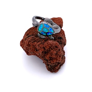 May include: A black ring with a blue opal turtle design. The ring is sitting on a piece of brown lava rock.