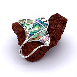May include: A silver pendant shaped like a manta ray with an iridescent abalone shell inlay. The pendant is sitting on a piece of brown lava rock.