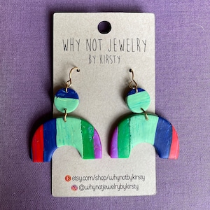 Può includere: Un paio di orecchini fatti a mano in argilla polimerica a forma di arcobaleno. Gli orecchini sono verdi, blu, rossi, viola e bianchi. Gli orecchini sono su una carta marrone con il testo "Why Not Jewelry by Kirsty" e le maniglie Etsy e Instagram del negozio.