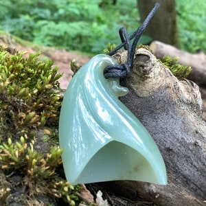 May include: A light green jade pendant shaped like a wave, hanging from a black cord. The pendant is carved with a smooth, flowing design and has a glossy finish.