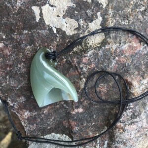 May include: A green jade pendant carved in the shape of a wave, hanging from a black cord.