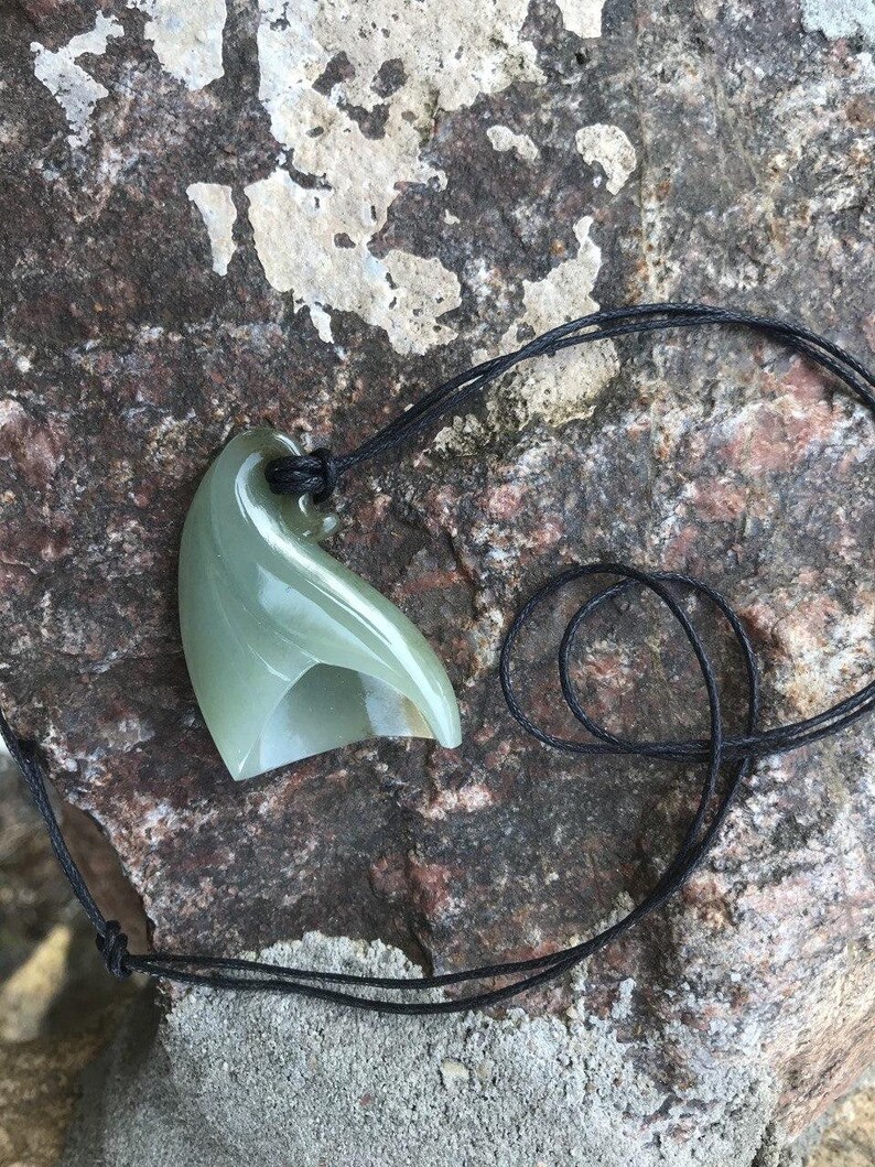 May include: A green jade pendant carved in the shape of a wave, hanging from a black cord.