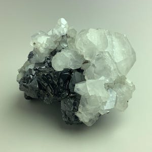 May include: A cluster of crystals featuring a mix of dark, metallic-looking minerals and translucent, clear quartz crystals. The quartz crystals have a faceted appearance, while the darker minerals have a more compact, irregular structure.