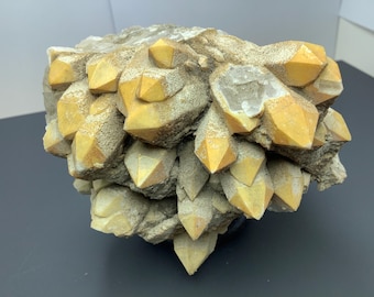 Può includere: Un gruppo di cristalli di quarzo con punte giallo dorato e una matrice grigia ruvida. I cristalli sono appuntiti e raggruppati, creando una suggestiva formazione naturale. La forma generale è irregolare e strutturata.