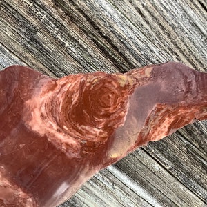 Puede incluir: Una piedra pulida de forma irregular con un patrón arremolinado de colores marrón rojizo y crema. La superficie de la piedra tiene una textura suave y descansa sobre una superficie de madera envejecida.