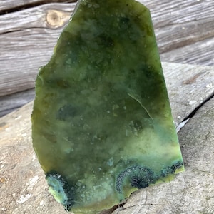May include: A large, irregularly shaped piece of green jade with dark green and black inclusions. The jade is polished and has a smooth surface.