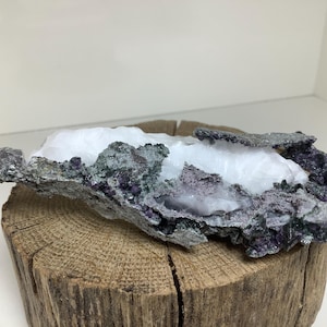 May include: A white crystal cluster with purple and green accents, resting on a wooden log. The crystal has a rough, natural texture.