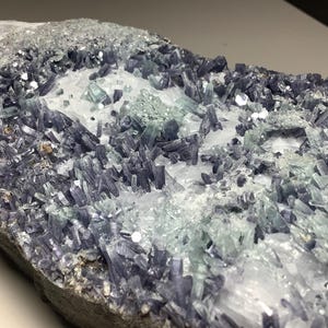 May include: A close-up of a mineral specimen featuring a matrix of gray rock with clusters of blue-gray, translucent crystals. The crystals are interspersed with areas of white and light green crystalline formations.