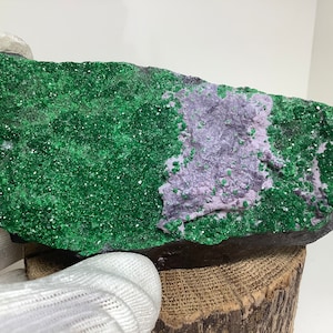 Pode incluir: Uma rocha grande, áspera, de cor cinza escura com um depósito mineral verde e roxo. O depósito mineral é composto por pequenos cristais brilhantes.