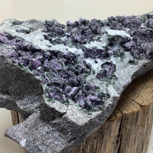 May include: A large, rough, gray rock with a cluster of purple and green crystals embedded in it. The rock is sitting on a piece of weathered wood.