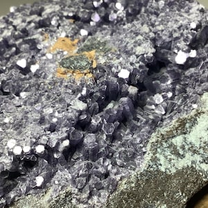 May include: A cluster of purple amethyst crystals on a dark gray rock. The crystals are small and have a sparkly, iridescent finish.