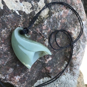 May include: A green jade pendant carved in the shape of a hook, hanging from a black cord. The pendant is smooth and polished, with a subtle sheen.