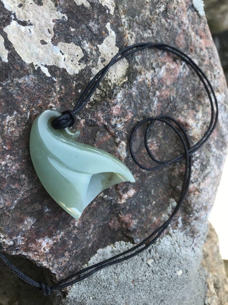 May include: A green jade pendant carved in the shape of a hook, hanging from a black cord. The pendant is smooth and polished, with a subtle sheen.