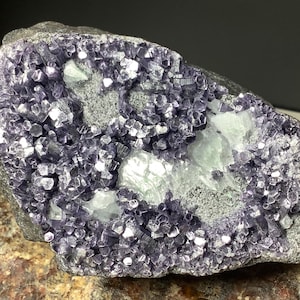 May include: A natural fluorite specimen with a mix of purple and white crystalline structures. The stone has a rough, textured surface with areas of gray and a dark background.