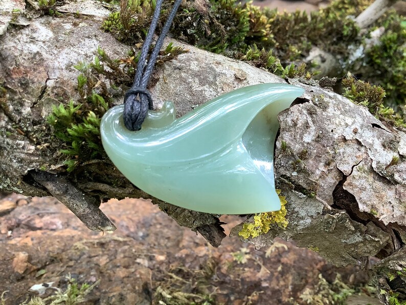May include: A pale green jade pendant shaped like a leaf, hanging from a black cord. The pendant is carved with intricate details and has a smooth, polished finish.